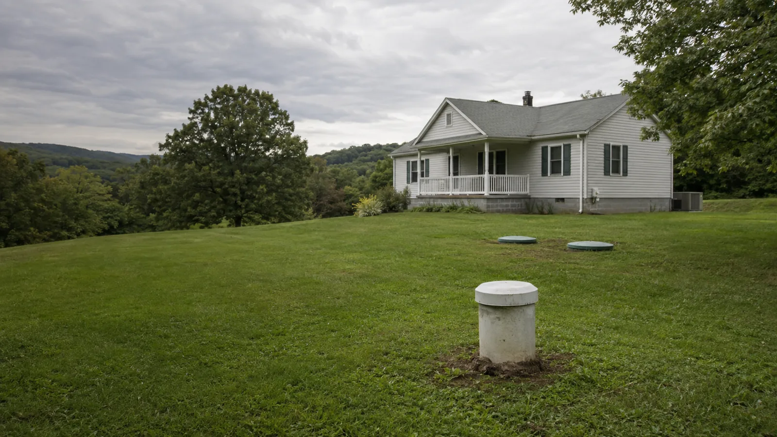 Illustrative view of a private well casing and septic access covers beside a house on rolling terrain
